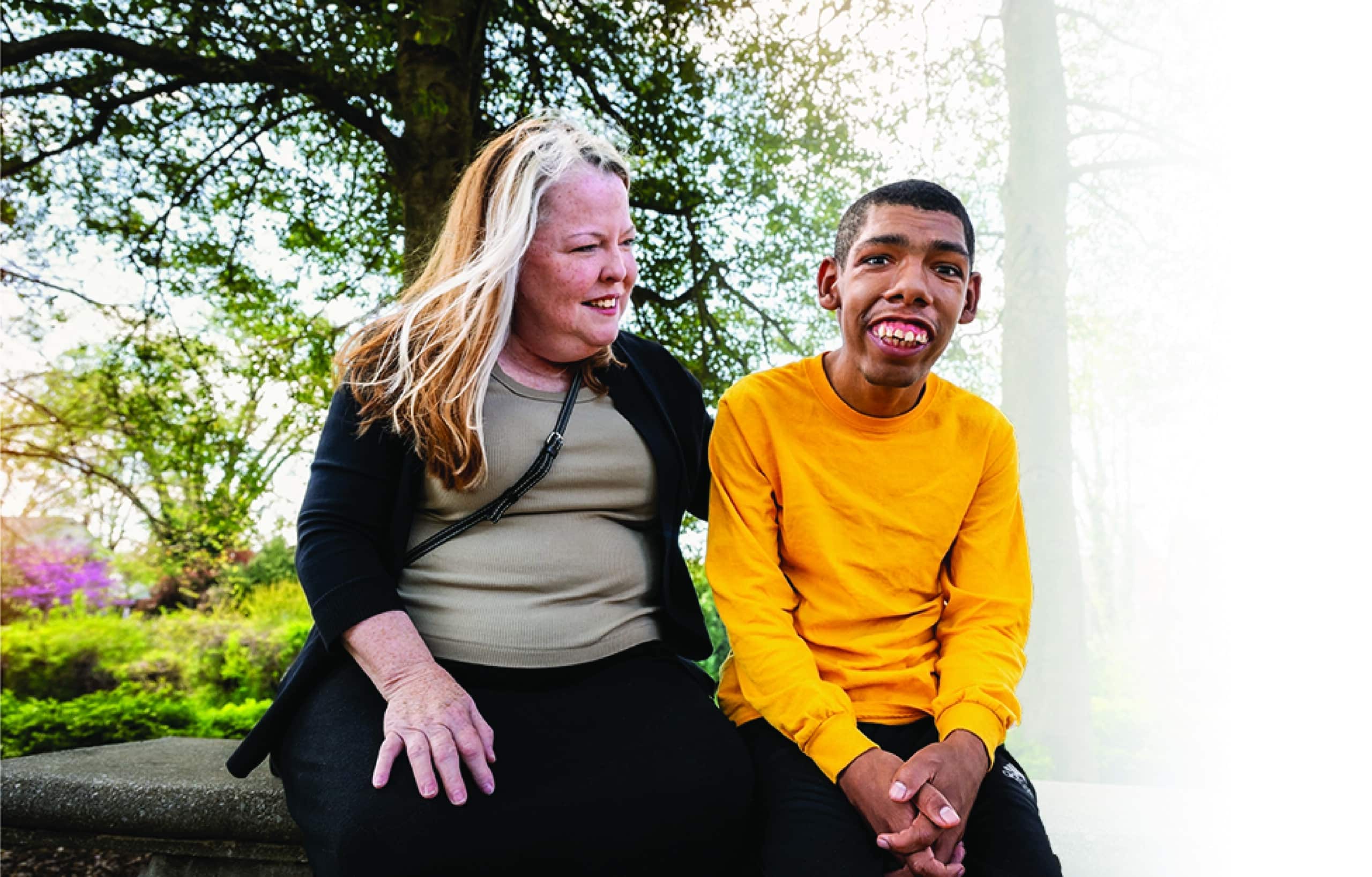 A woman smiling and sitting beside a young man with a disability, both outdoors under a tree on a sunny day.