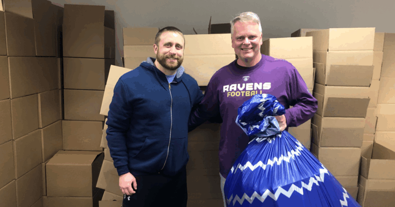 Two men standing in front of empty boxes holding a bag of Christmas gifts