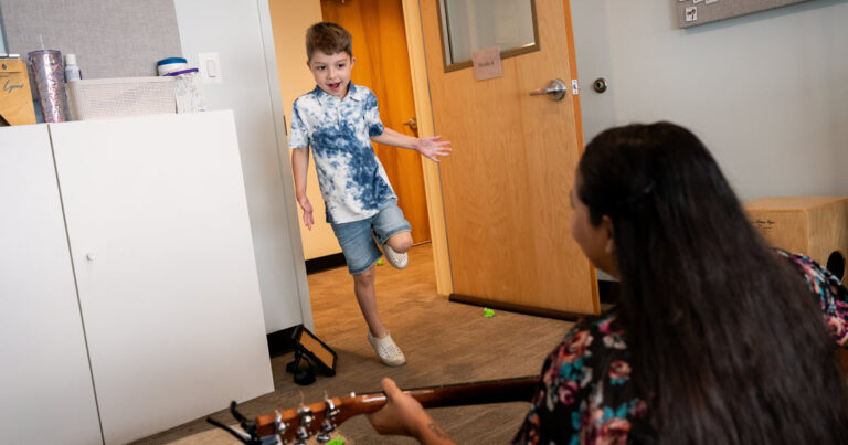 Little boy jumping through a door frame excited to be going to music therapy. Music teacher in the foreground holding a guitar
