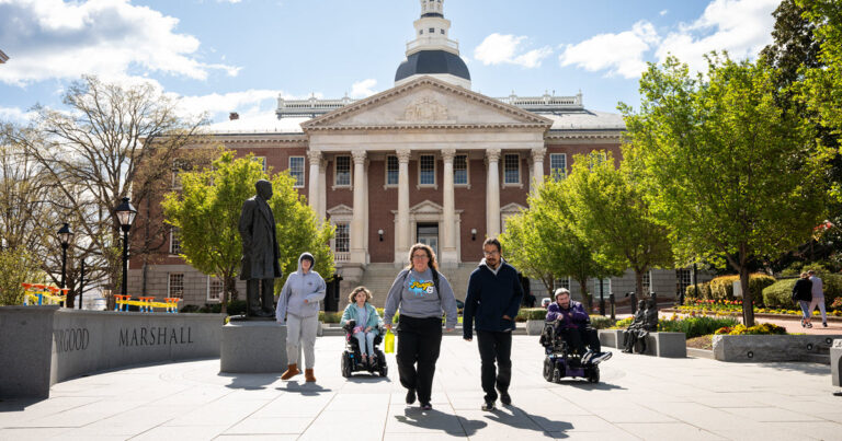 Five people, 3 walking and 2 in wheelchairs on Lawyers Mall in front of the Maryland State Capital building.