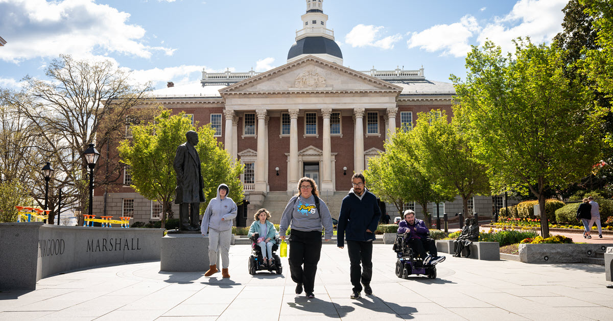 Five people, 3 walking and 2 in wheelchairs on Lawyers Mall in front of the Maryland State Capital building.