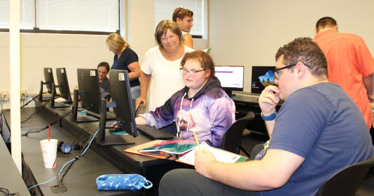 Two students in a classroom working on a computer while teacher helps