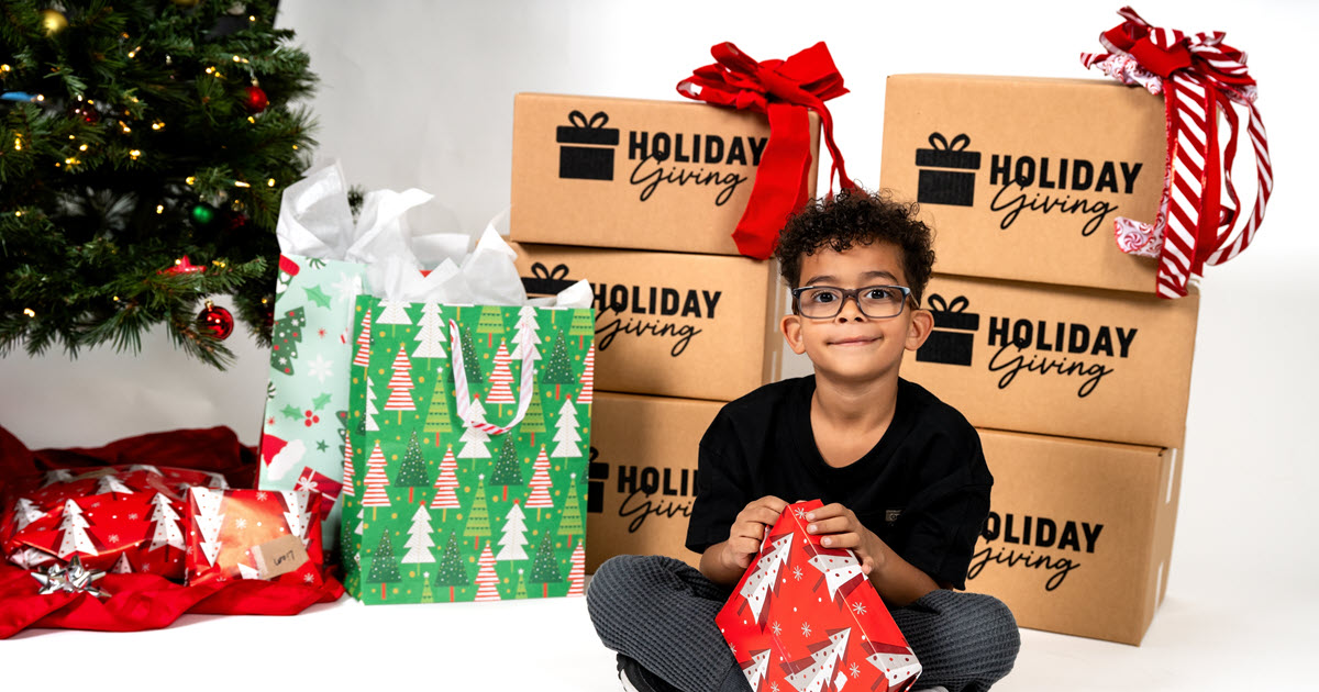 A boy sitting in front of a Christmas tree with gifts and boxes that say Holiday Giving on them