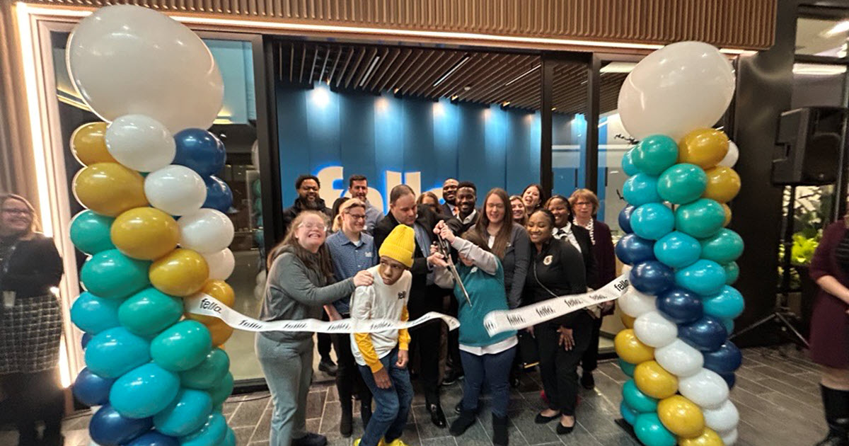 A group of people celebrating a ribbon-cutting ceremony indoors for Fello at The Mall, flanked by two balloon columns with blue, white, and yellow balloons.