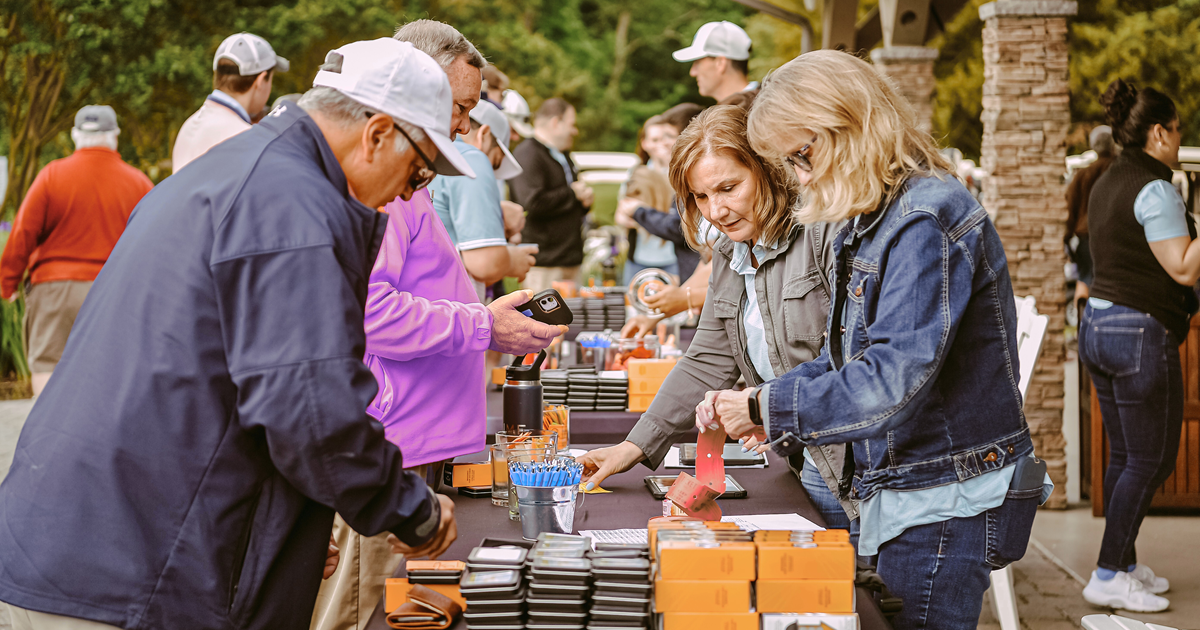 Men are buying 50/50 raffle tickets from two women at a table stacked with golf balls and golf gifts