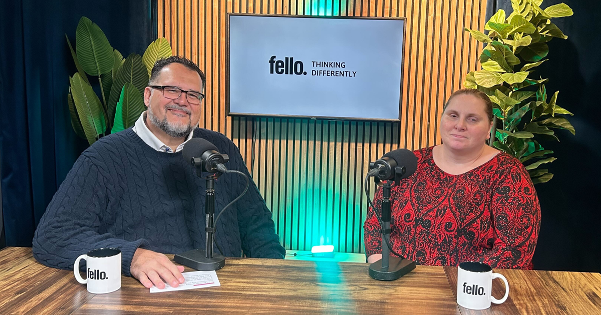 Two individuals are seated at a podcast table with microphones. Behind them, a screen displays the text "fello. THINKING DIFFERENTLY." The table holds two coffee mugs, both labeled "fello." The setting includes green plants and wood paneling.