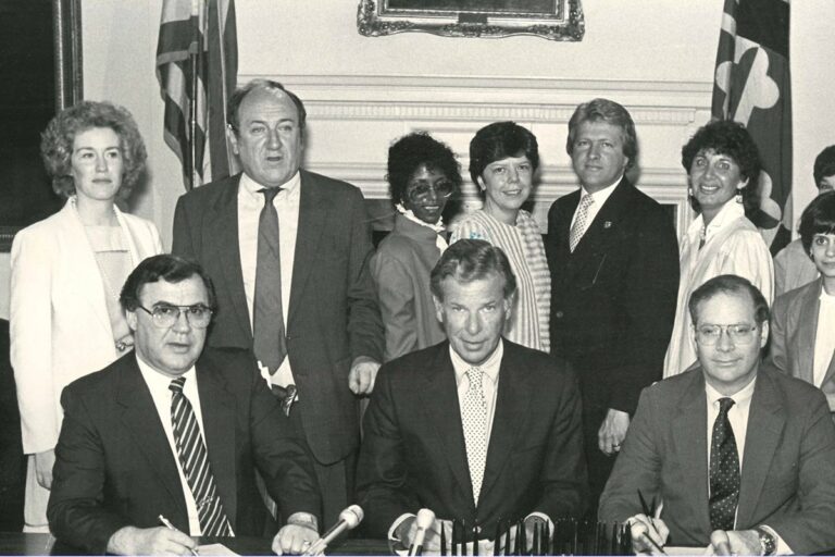 A group of people, some seated and others standing, are gathered in a formal setting. Three people are seated at a table with microphones, likely involved in an official event or signing. Several flags are visible in the background. The scene suggests an important occasion or announcement.