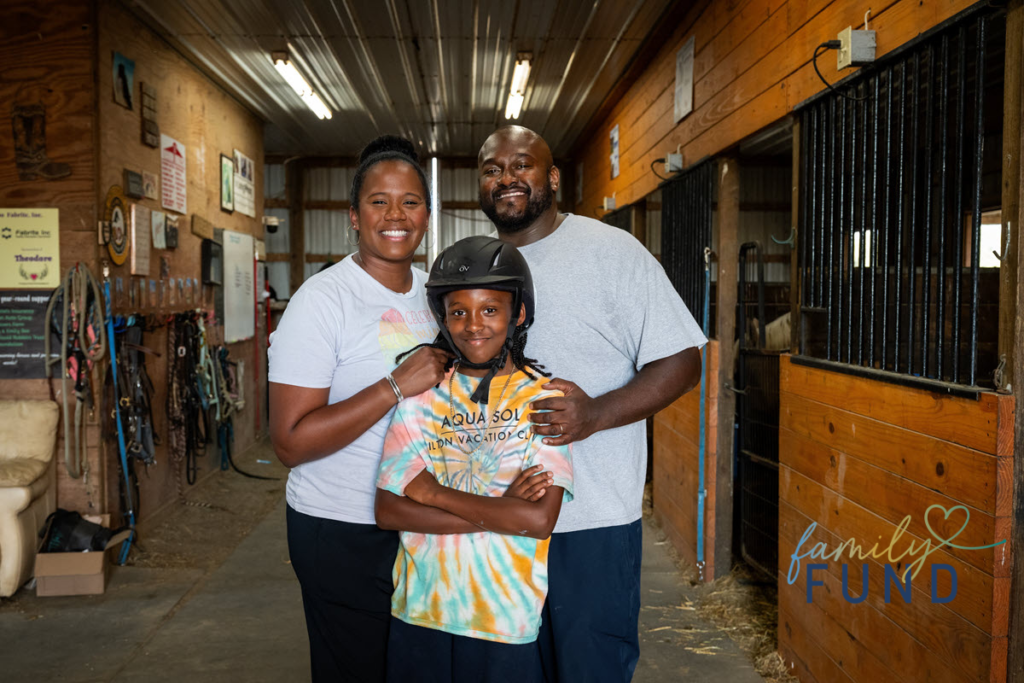 Three people standing in a horse barn smiling for the camera. The young man in the middle is wearing a helmet
