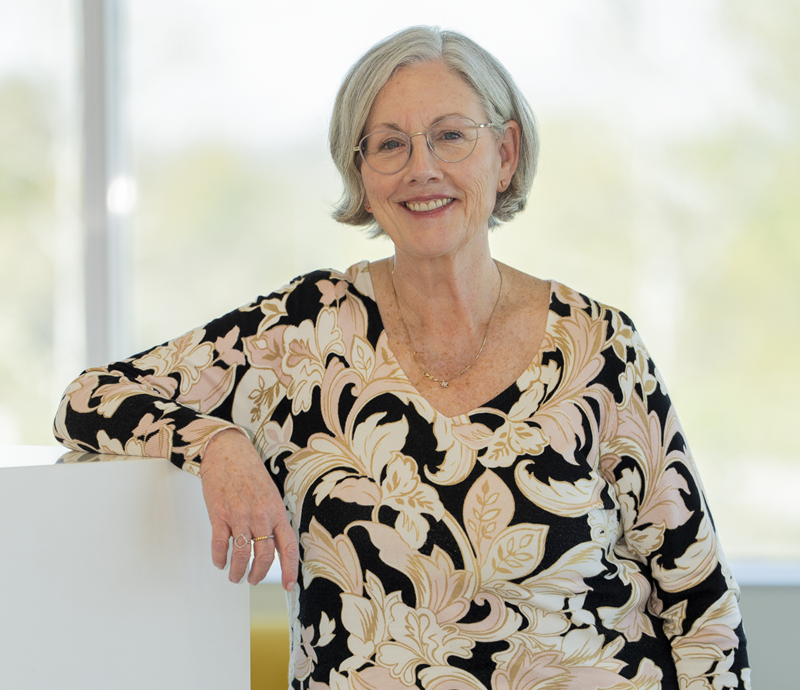 Meg Hayes headshot. She's in a bright well-lit room with white hair, wearing glasses and a black and gold floral shirt