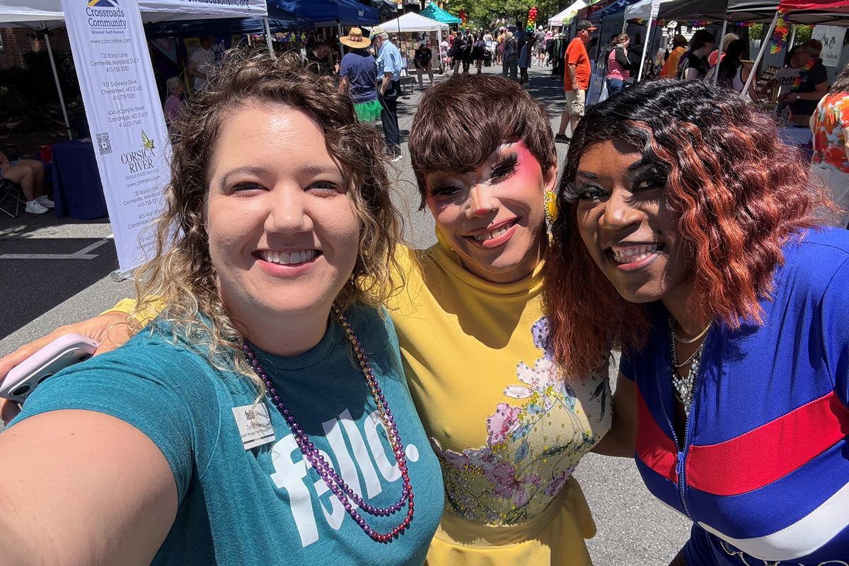Three people smiling outdoors at a lively street festival with tents and vendors in the background. One person is wearing a teal shirt, another a yellow outfit with floral designs, and the third a blue, red, and white outfit.
