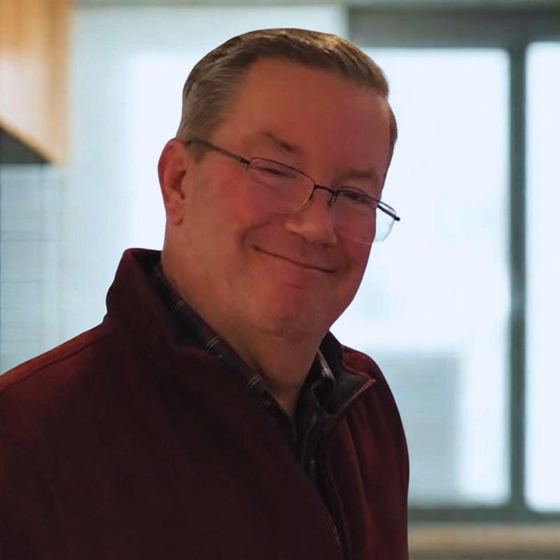 Steve Brennan smiling for the camera wearing glasses and a red sweater. 