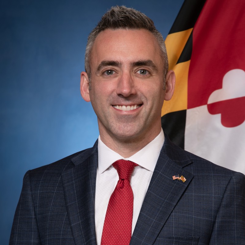 Jake Day, Secretary, Department of Housing & Community Development stand in front of the Maryland Flag wearing a blue suit and red tie. 