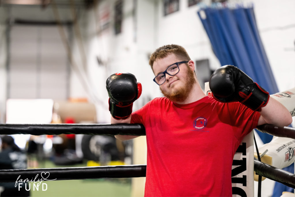 A person wearing a red t-shirt, boxing gloves and glasses while standing in the corner of a boxing ring