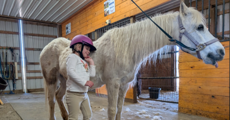 A little girl leaning her head on a horse wearing riding gear and a pink helmet